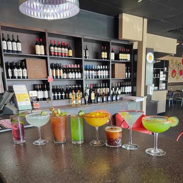 A bar counter at Ripe Eatery with seven colorful cocktails and wine bottles on shelves in the background.