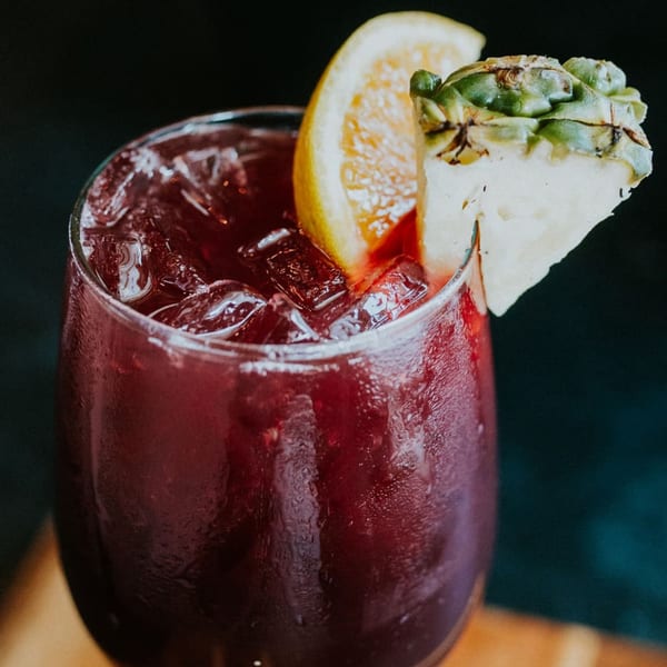 Close-up of a red drink with ice, garnished with an orange slice and pineapple wedge.