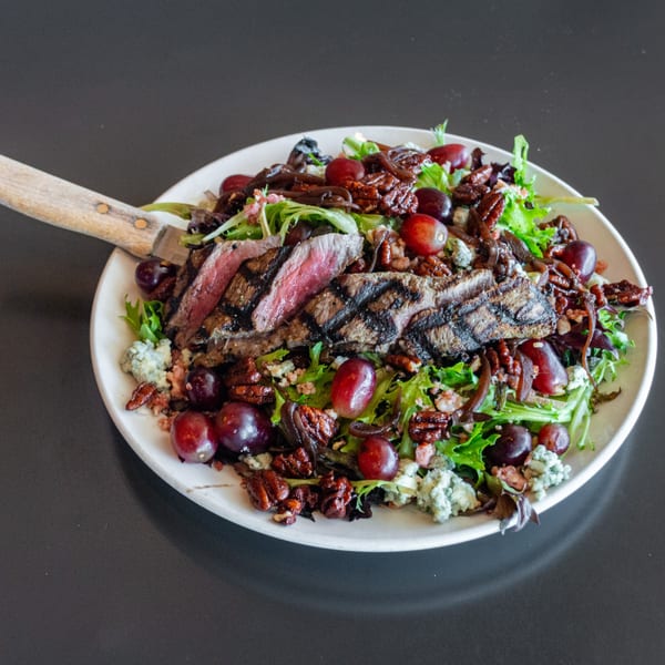 Grilled steak salad with grapes, pecans, blue cheese, and mixed greens on a white plate.