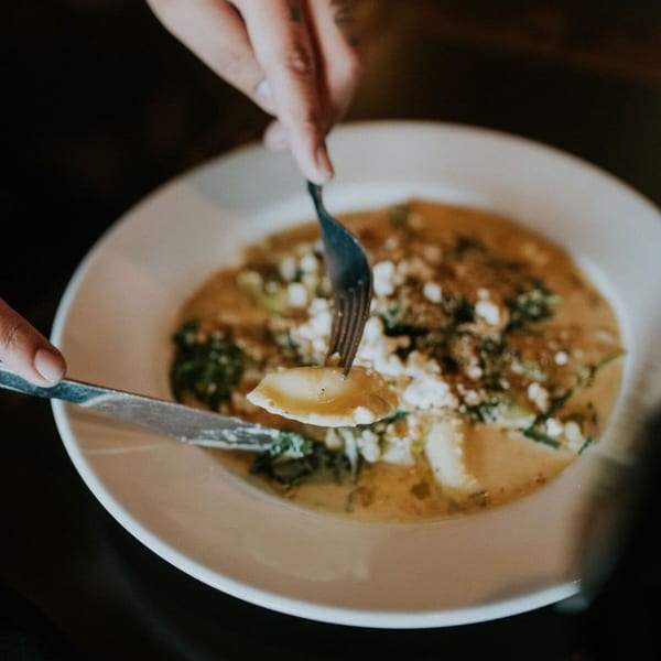 A person cutting a piece of ravioli with a fork and knife from a creamy dish.