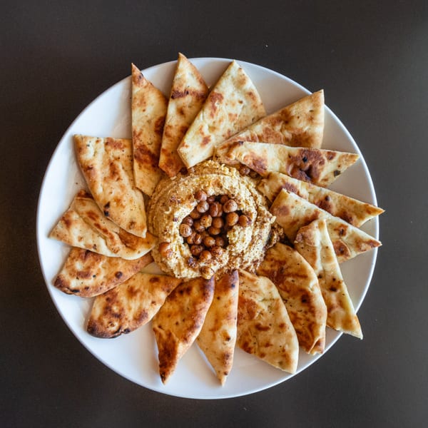 A white plate of hummus topped with chickpeas, surrounded by toasted pita bread triangles.