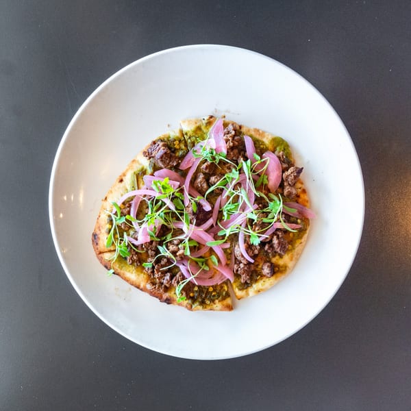 Flatbread with ground meat, green sauce, pickled red onions, and microgreens on a white plate.