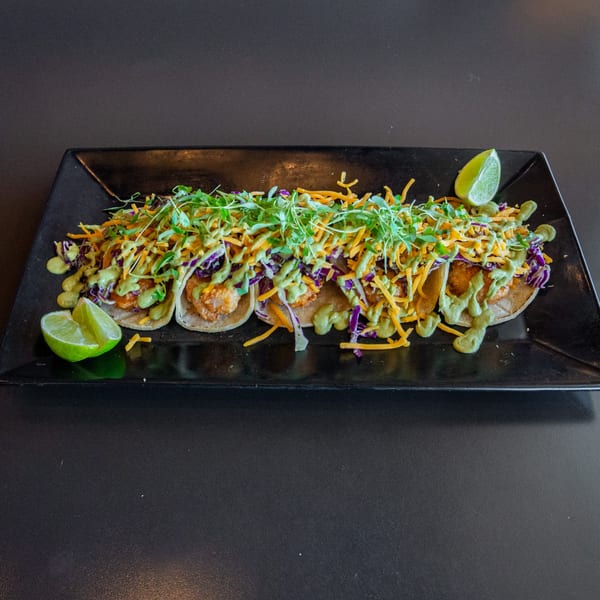 A black rectangular plate holds five shrimp tacos topped with shredded cheese, cabbage, and green sauce.