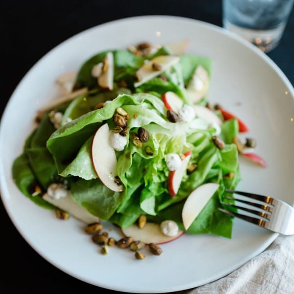 A fresh green salad with apple slices, pistachios, and goat cheese on a white plate.