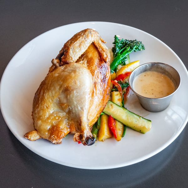 Golden-brown roasted chicken with broccolini, zucchini, bell peppers, and a side of sauce on a white plate.