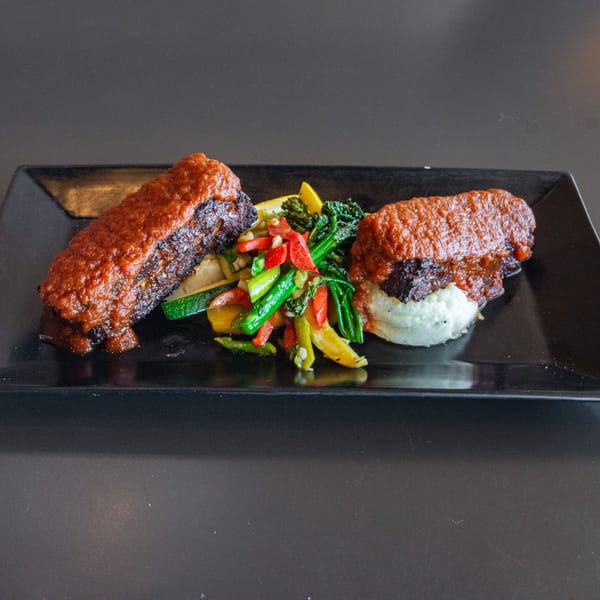 Two ribs with red sauce, mashed potatoes, and mixed vegetables on a black rectangular plate.
