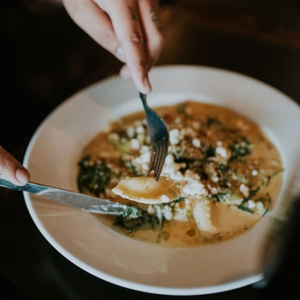 Hands using a fork and knife to eat ravioli in a creamy sauce with greens and cheese.